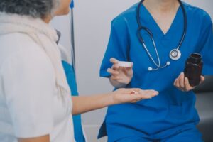 Asian nurse explains medication to senior woman before administering. Home caregiver visit ensures proper care and medication guidance for elderly patients.