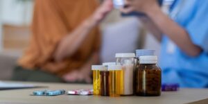 Close-up of medication bottles and pills on a table, with an elderly woman and caregiver in the background. Elderly care and medication concept.