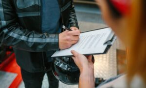 A biker man wearing leather jacket and holding helmet signing insurance policy to receipt his repaired motorcycle on workshop