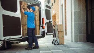 Delivery Man Uses Hand Truck Trolley Full of Cardboard Boxes and Packages, Loads Parcels into Truck / Van. 